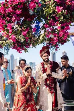 A happy North Indian bride and groom standing together in traditional wedding attire under a floral mandap
